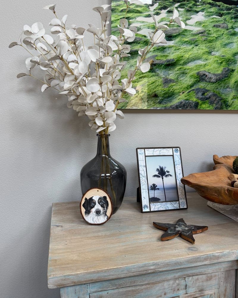 Hand-painted black-and-white collie puppy on a wood round ornament, resting against a dark glass vase of white eucalyptus
