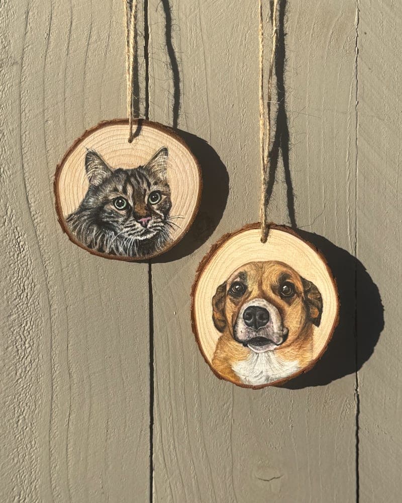 Two hand-painted wood round ornaments hanging by twine on a fence — a brown tabby cat and a tan-and-white dog