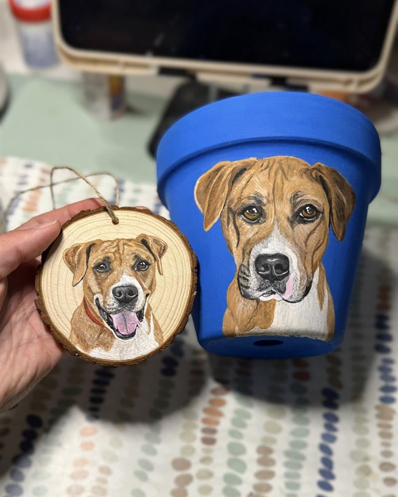 Hand-painted dog on a wood round ornament beside a matching dog portrait on a blue pot